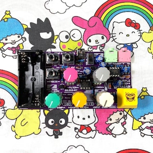 May include: A purple circuit board with various coloured knobs and jacks, featuring the text "DIG.DUG.DIY" and "PURPLE AIN AGAIN". The board is set against a colourful cartoon character patterned background.