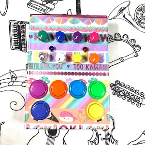 May include: A decorative box covered in colourful washi tape, featuring various colourful magnets and push pins. The box is adorned with text that reads "KAWAII" and "ALL FOR YOU TOO KAWAII!!" The magnets are in shades of purple, orange, blue, and green.