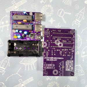 Peut inclure: Une carte de circuit imprimé USB2USB MIDI HOST violette avec des composants électroniques, dont des puces, des résistances et des ports USB. Un support de batterie noir et une deuxième carte de circuit imprimé violette sont également visibles. Le texte comprend "DIG.DUG.DIY".