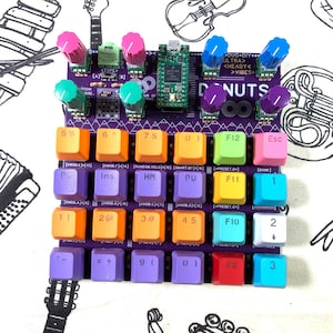 May include: A purple electronic music device with colorful knobs and keys. The device has a grid of multi-colored keys, labeled with numbers, symbols, and function keys. The top has several knobs in various colors, and the text "DONUTS" is visible.