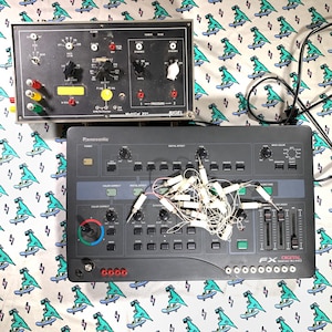 May include: A black Rigel MultiCal 201 medical calibration device with knobs and ports positioned above a black Panasonic WJ-AVE5 digital video mixer featuring numerous buttons, sliders, and a tangled cluster of white cables resting on the surface.