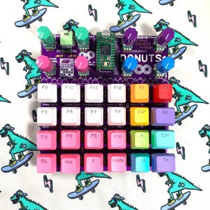Pode incluir: Um controlador de música eletrónica roxo com uma grelha de teclas coloridas etiquetadas com funções como F1 a F12, Alt e Ctrl. O dispositivo inclui vários botões rotativos em menta, rosa, roxo e azul, juntamente com uma placa de circuito.