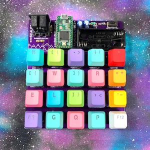 May include: A DIY electronic music synthesizer with colorful square keys. The keys are arranged in a grid on a purple circuit board. The board includes text that reads "DIG DUG DIY" and "TRA.HEADY.VIBES".