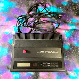 May include: A black Yamaha REX50 digital multi-effector unit featuring a rectangular chassis, a small LCD screen, a series of control buttons, and a single red knob. A black power cord with a two-prong plug is coiled on top of the device.