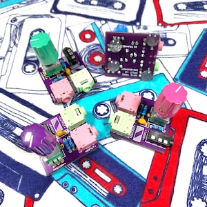 Puede incluir: Una colección de placas de circuito púrpura con perillas de colores y conectores de entrada/salida. Las placas tienen texto que dice "GAIN", "INPUT", "OUTPUT" y "TRRS". El fondo es una tela con un patrón de cinta de cassette.