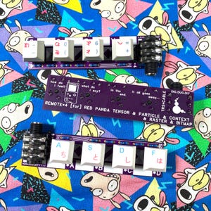 May include: Two purple electronic circuit boards with white keycaps and black jacks. One board has the text "REMOTE+4 [for] RED PANDA TENSOR & PARTICLE & CONTEXT & RASTER & BITMAP". The other board has Japanese characters and numbers. The background is a colorful cartoon print.