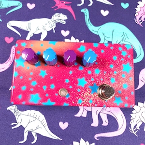 May include: A rectangular guitar effects pedal with a vibrant pink and orange gradient finish, decorated with bright cyan stars and gold glitter. It features four ridged control knobs in alternating purple and blue, a small circular LED, and a metal footswitch.