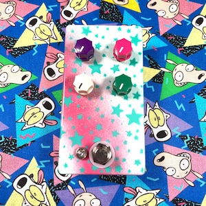 May include: A rectangular guitar effects pedal featuring a white and pink gradient finish with teal star patterns. The device includes four colorful control knobs in purple, pink, white, and green, along with a silver metal footswitch and a small indicator light.