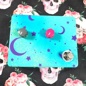 Puede incluir: Un pedal de efectos para guitarra rectangular con un acabado turquesa brillante que presenta lunas crecientes y estrellas de color púrpura. El pedal incluye una perilla gris, una perilla rosa y un interruptor de pie metálico plateado.
