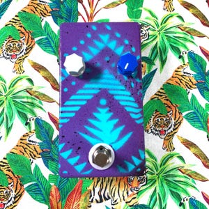 Peut inclure: Une pédale d'effet pour guitare rectangulaire avec une base violette ornée d'un motif géométrique cyan vif et de taches noires. L'appareil comprend un bouton blanc, un bouton bleu et un commutateur au pied en métal argenté en bas.
