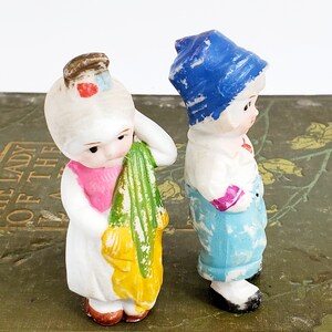 Puede incluir: Dos figuras antiguas de porcelana de ni&ntilde;os. Una figura viste un vestido blanco con cuello rosa y sostiene una tela verde y amarilla. La otra figura lleva un gorro azul, camisa blanca y pantal&oacute;n azul. Ambas tienen rasgos faciales pintados.