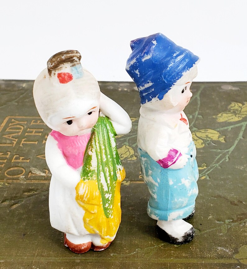 Puede incluir: Dos figuras antiguas de porcelana de ni&ntilde;os. Una figura viste un vestido blanco con cuello rosa y sostiene una tela verde y amarilla. La otra figura lleva un gorro azul, camisa blanca y pantal&oacute;n azul. Ambas tienen rasgos faciales pintados.