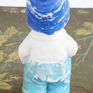 Puede incluir: Peque&ntilde;a figura vintage de una persona con sombrero azul, camisa blanca y pantalones cortos azules. La figura est&aacute; pintada con un acabado envejecido. El texto "MADE IN JAPAN" y "CA/760" son visibles en la parte posterior. Sobre una superficie verde.