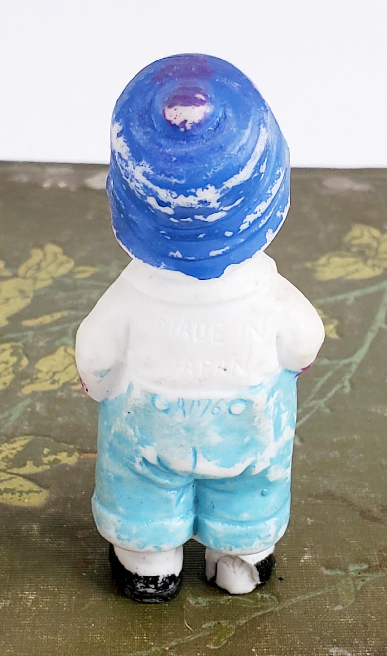 Puede incluir: Peque&ntilde;a figura vintage de una persona con sombrero azul, camisa blanca y pantalones cortos azules. La figura est&aacute; pintada con un acabado envejecido. El texto "MADE IN JAPAN" y "CA/760" son visibles en la parte posterior. Sobre una superficie verde.