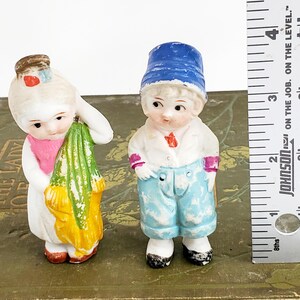 Puede incluir: Dos figuras antiguas de porcelana. Una es una ni&ntilde;a con un vestido blanco con una bufanda rosa y un objeto verde y amarillo. El otro es un ni&ntilde;o con un sombrero azul, camisa blanca y pantalones azules. Una regla est&aacute; al lado de las figuras.