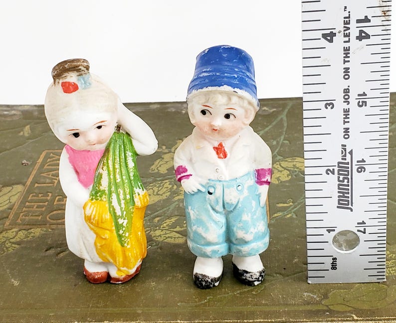 Puede incluir: Dos figuras antiguas de porcelana. Una es una ni&ntilde;a con un vestido blanco con una bufanda rosa y un objeto verde y amarillo. El otro es un ni&ntilde;o con un sombrero azul, camisa blanca y pantalones azules. Una regla est&aacute; al lado de las figuras.
