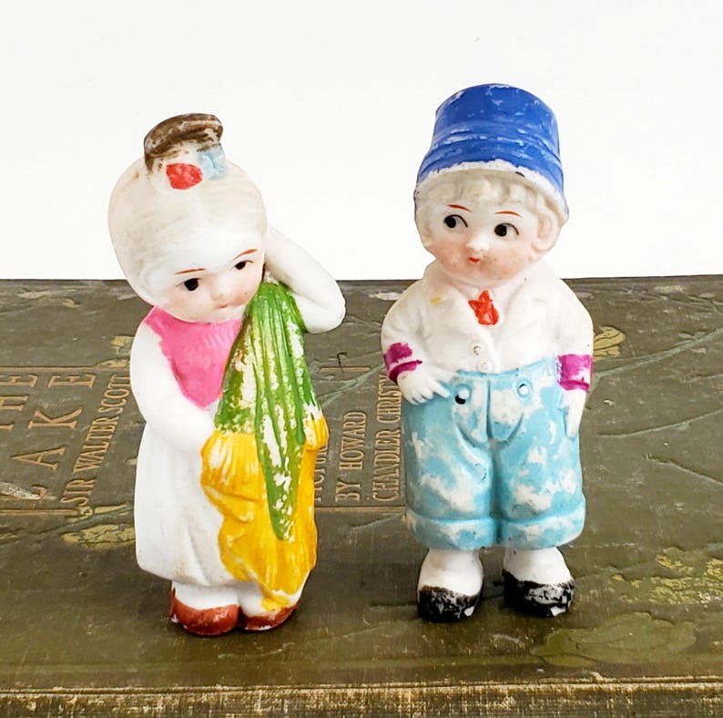 Puede incluir: Dos peque&ntilde;as figuras de porcelana antiguas. Una es una ni&ntilde;a con un vestido blanco con una parte superior rosa y un chal verde y amarillo. El otro es un ni&ntilde;o con un sombrero azul, una chaqueta blanca y pantalones cortos azules. Ambos tienen detalles pintados.