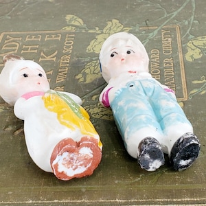 Puede incluir: Dos mu&ntilde;ecas de porcelana vintage. Una mu&ntilde;eca viste un atuendo blanco con detalles amarillos y verdes. La otra mu&ntilde;eca lleva un peto azul y una camisa blanca. Ambas mu&ntilde;ecas tienen rasgos faciales pintados.