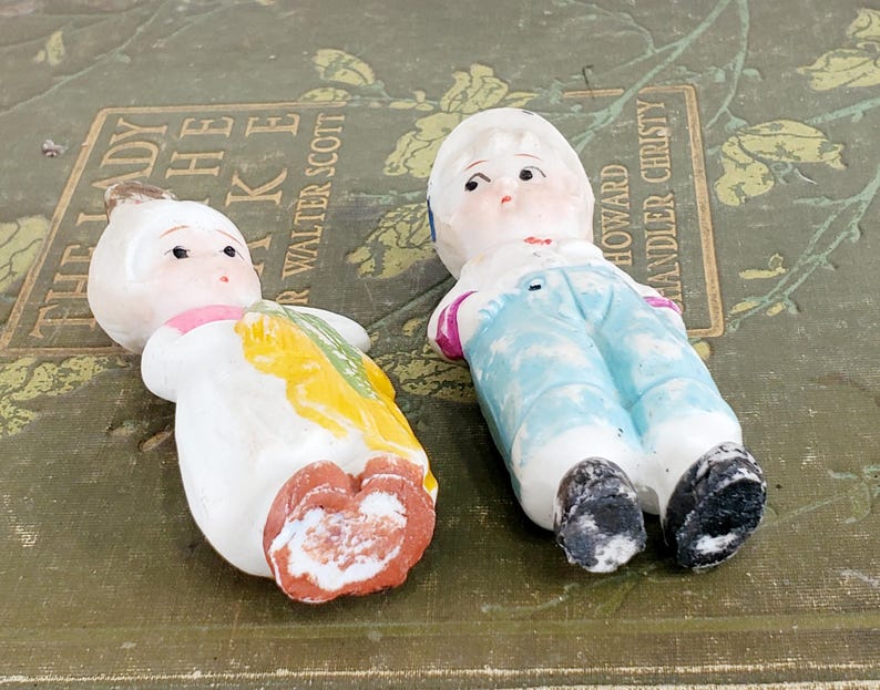 Puede incluir: Dos mu&ntilde;ecas de porcelana vintage. Una mu&ntilde;eca viste un atuendo blanco con detalles amarillos y verdes. La otra mu&ntilde;eca lleva un peto azul y una camisa blanca. Ambas mu&ntilde;ecas tienen rasgos faciales pintados.