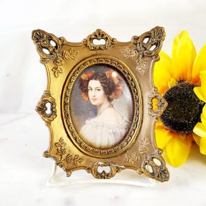May include: Ornate gold-colored oval picture frame with a portrait of a woman in a white dress and a sunflower in the background. The frame has a decorative design and a small stand.
