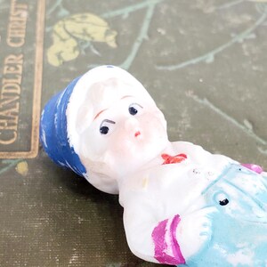 Puede incluir: Una peque&ntilde;a mu&ntilde;eca de porcelana vintage con una cara pintada y un sombrero azul. La mu&ntilde;eca lleva una camisa blanca con un lazo rojo y un peto azul claro con rayas rosas. La mu&ntilde;eca est&aacute; tumbada sobre una superficie verde.