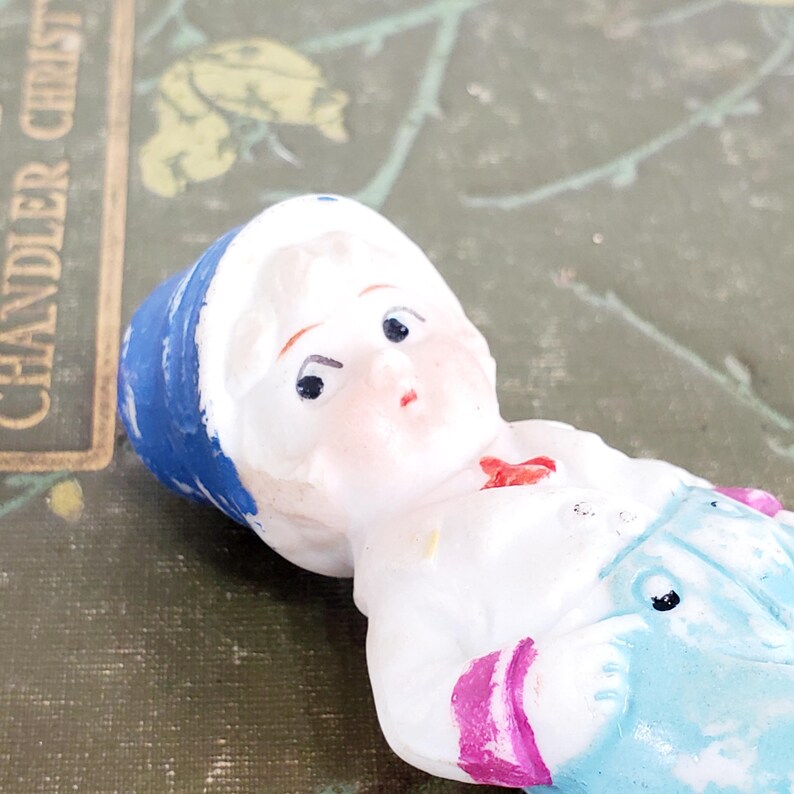 Puede incluir: Una peque&ntilde;a mu&ntilde;eca de porcelana vintage con una cara pintada y un sombrero azul. La mu&ntilde;eca lleva una camisa blanca con un lazo rojo y un peto azul claro con rayas rosas. La mu&ntilde;eca est&aacute; tumbada sobre una superficie verde.