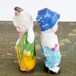 Puede incluir: Dos figuras de cer&aacute;mica vintage de ni&ntilde;os. Una lleva un gorro blanco, un lazo rosa, una falda amarilla y un chal verde. La otra lleva un gorro azul, una camisa blanca y pantalones azules. Ambos est&aacute;n de espaldas.