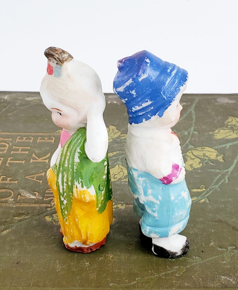 Puede incluir: Dos figuras de cer&aacute;mica vintage de ni&ntilde;os. Una lleva un gorro blanco, un lazo rosa, una falda amarilla y un chal verde. La otra lleva un gorro azul, una camisa blanca y pantalones azules. Ambos est&aacute;n de espaldas.