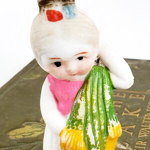 Puede incluir: Figura de porcelana vintage de una ni&ntilde;a sosteniendo una tela verde y amarilla. La ni&ntilde;a tiene un vestido blanco, una bufanda rosa y un sombrero marr&oacute;n con detalles rojos y azules. La figura est&aacute; sobre un libro.