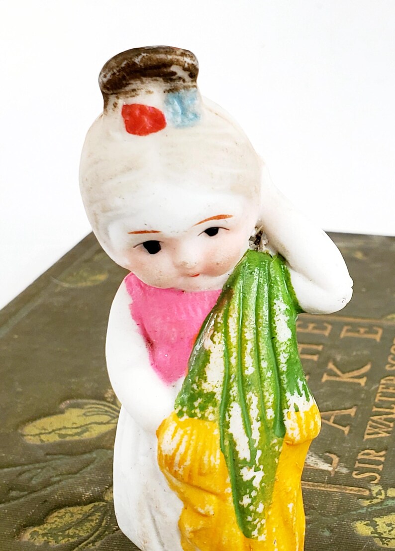 Puede incluir: Figura de porcelana vintage de una ni&ntilde;a sosteniendo una tela verde y amarilla. La ni&ntilde;a tiene un vestido blanco, una bufanda rosa y un sombrero marr&oacute;n con detalles rojos y azules. La figura est&aacute; sobre un libro.