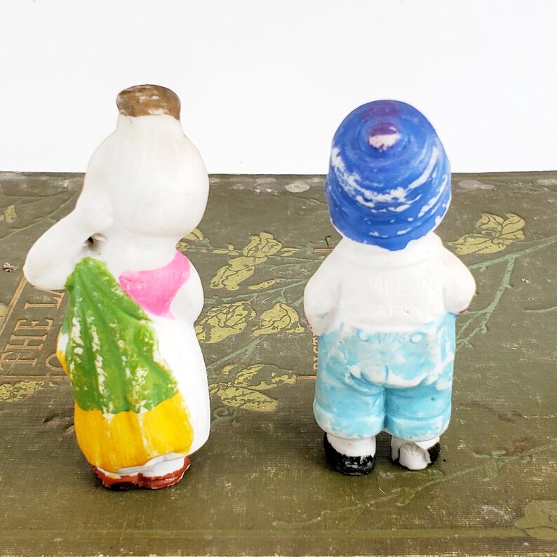 Puede incluir: Dos peque&ntilde;as figuras de porcelana vintage. Una figura lleva un vestido blanco con un top rosa y sostiene un objeto verde y amarillo. La otra figura lleva una camisa blanca, pantalones cortos azules y un sombrero azul. Ambas figuras tienen detalles pintados.