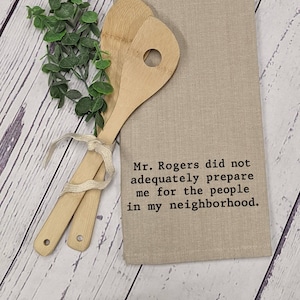 May include: A beige kitchen towel with the text "Mr. Rogers did not adequately prepare me for the people in my neighborhood." embroidered in black. Two wooden spoons are tied together with a white ribbon and are lying on a white wooden surface.