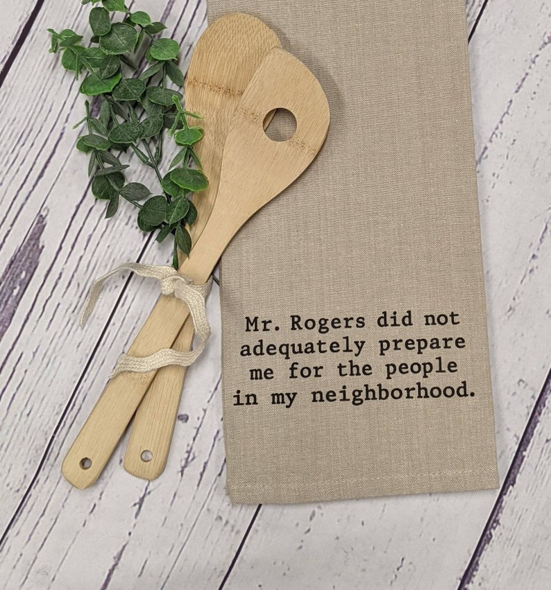 May include: A beige kitchen towel with the text "Mr. Rogers did not adequately prepare me for the people in my neighborhood." embroidered in black. Two wooden spoons are tied together with a white ribbon and are lying on a white wooden surface.
