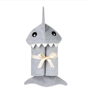 May include: A gray hooded towel shaped like a shark with a white mouth and black eyes. The towel is tied with a cream-colored ribbon.