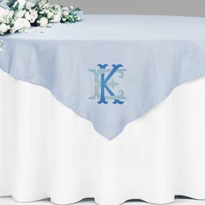 May include: A light blue table overlay with a white table underneath. The overlay has a blue and white monogram of the letter "K" in the center.