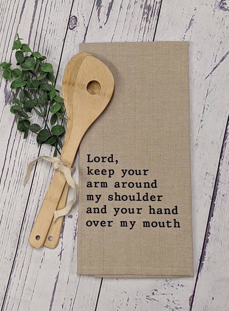 May include: A beige kitchen towel with the text "Lord, keep your arm around my shoulder and your hand over my mouth" printed in black.