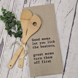 May include: A beige kitchen towel with the text "Good moms let you lick the beaters, great moms turn them off first" printed in black. Two wooden spoons are placed on top of the towel.