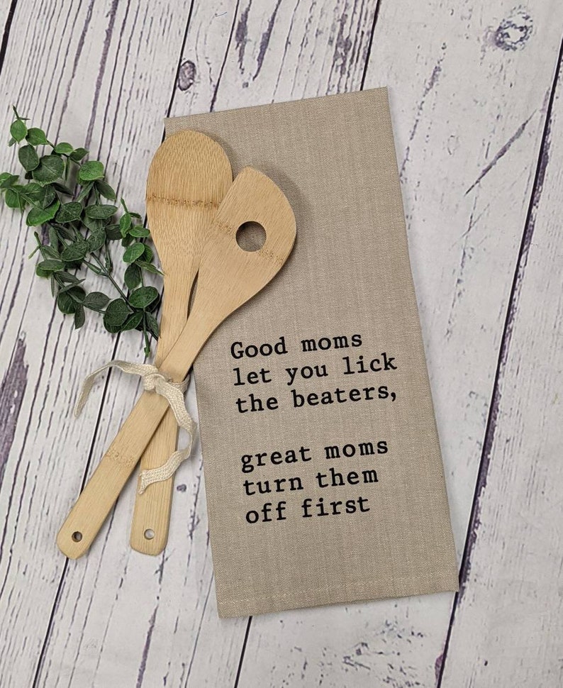May include: A beige kitchen towel with the text "Good moms let you lick the beaters, great moms turn them off first" printed in black. Two wooden spoons are placed on top of the towel.