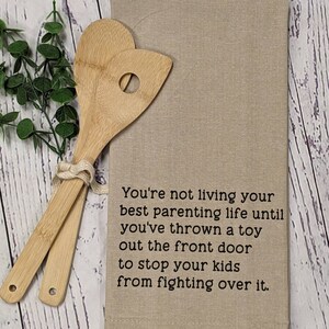 May include: A beige kitchen towel with the text "You're not living your best parenting life until you've thrown a toy out the front door to stop your kids from fighting over it."