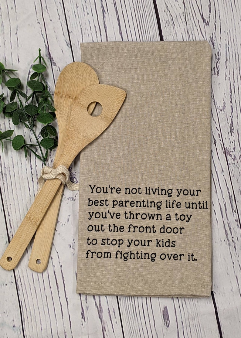 May include: A beige kitchen towel with the text "You're not living your best parenting life until you've thrown a toy out the front door to stop your kids from fighting over it."