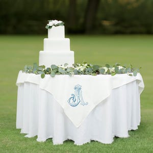 May include: A white wedding cake with a floral topper sits on a white table with a white linen tablecloth featuring a blue embroidered letter "A".