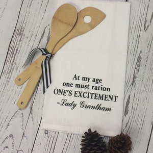 May include: A white kitchen towel with the quote "At my age one must ration ONE'S EXCITEMENT -Lady Grantham" in black script. Two wooden spoons are tied with a black and white striped ribbon. Two pinecones are also present.