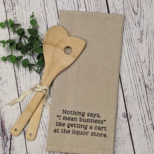 May include: Beige kitchen towel with the text "Nothing says, "I mean business" like getting a cart at the liquor store."  Two wooden spoons are on top of the towel.