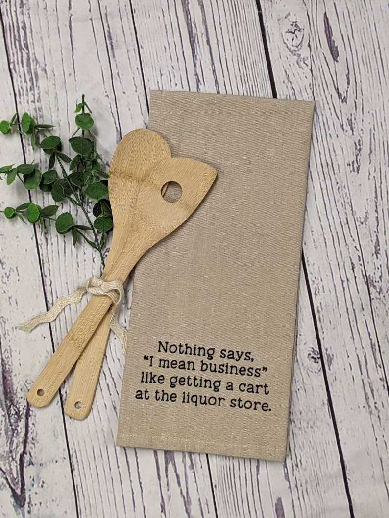 May include: Beige kitchen towel with the text "Nothing says, "I mean business" like getting a cart at the liquor store."  Two wooden spoons are on top of the towel.