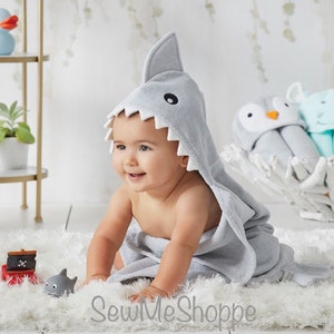 May include: A baby wearing a gray hooded towel shaped like a shark. The towel has a white trim and black eyes. The baby is sitting on a white fluffy rug.