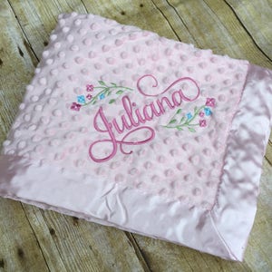 May include: Pink baby blanket with a satin border and embroidered name "Juliana" with floral accents.