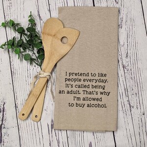 May include: A beige kitchen towel with the text "I pretend to like people everyday. It's called being an adult. That's why I'm allowed to buy alcohol."