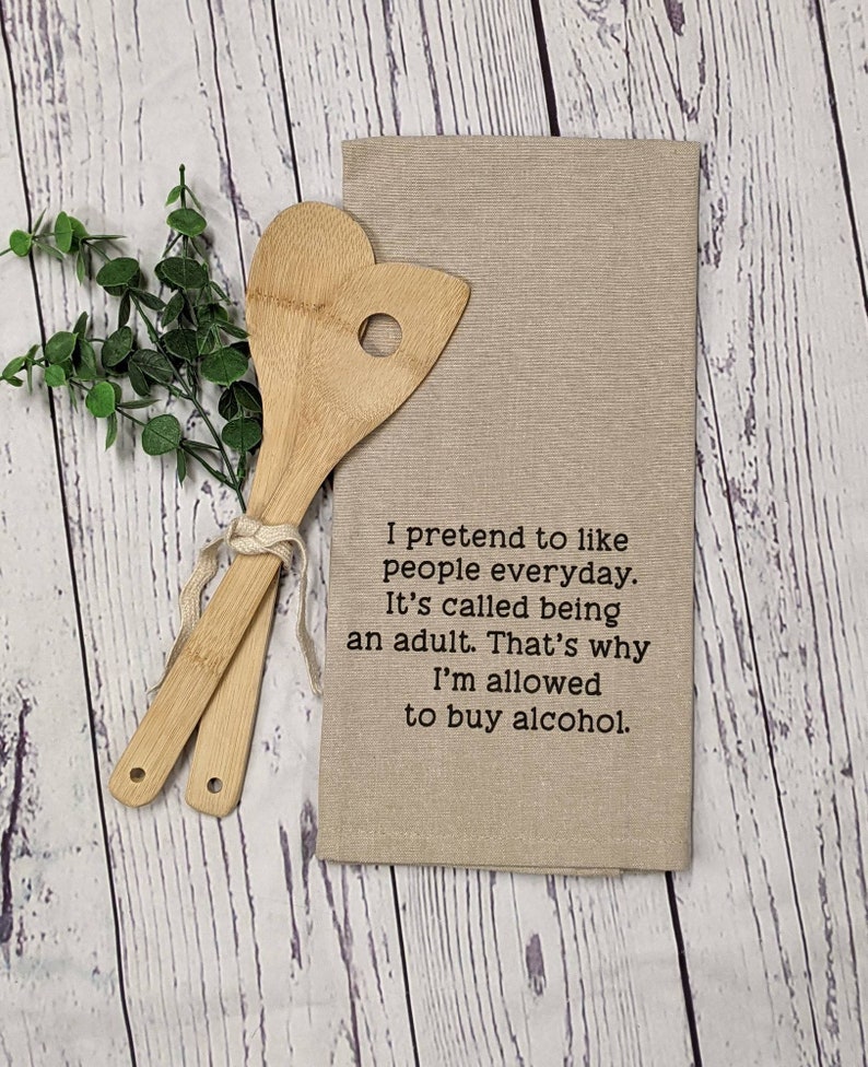 May include: A beige kitchen towel with the text "I pretend to like people everyday. It's called being an adult. That's why I'm allowed to buy alcohol."