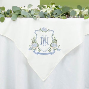 May include: White linen table runner with a blue and white floral crest featuring the monogram "NW" and the text "August 5, 2023".