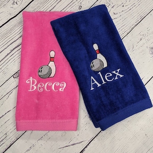 May include: Two personalized towels with a bowling pin and ball design. The towel on the left is pink with the name "Becca" embroidered on it. The towel on the right is blue with the name "Alex" embroidered on it.